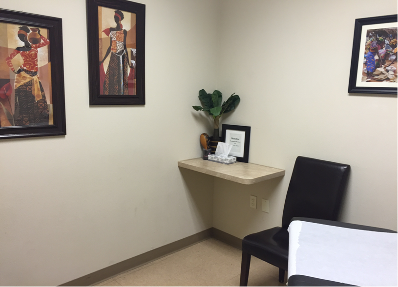 MomDoc Westridge OB-GYN exam room with framed African-figure prints, corner wood shelf with plant, and leather chair
